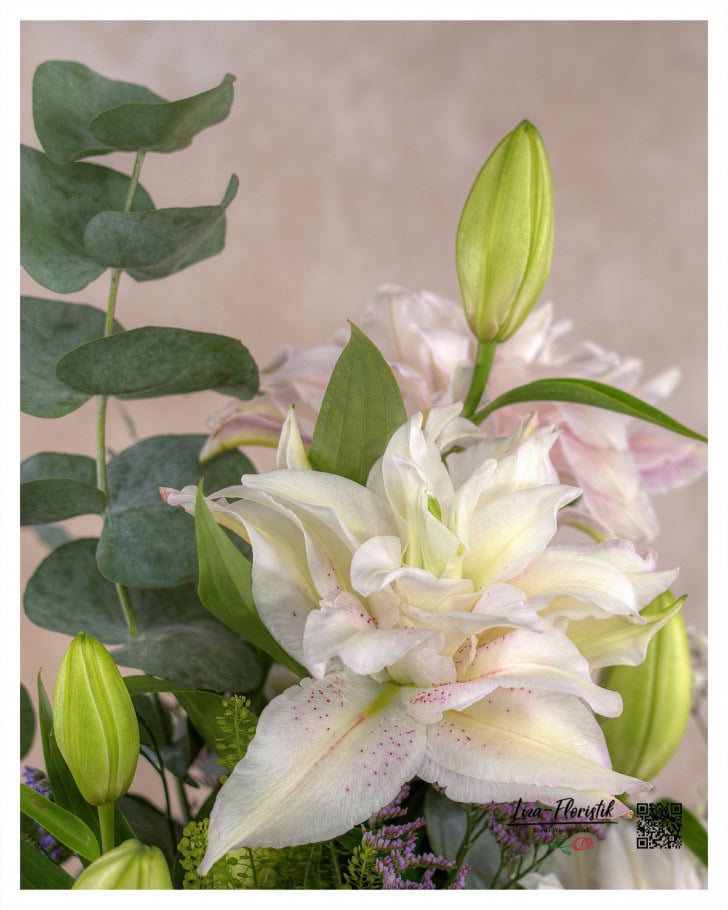 Eine zarte, gefüllte Lilie in Cremeweiß steht im Mittelpunkt des Trauergestecks, umgeben von Eukalyptusblättern und weiteren floralen Akzenten.