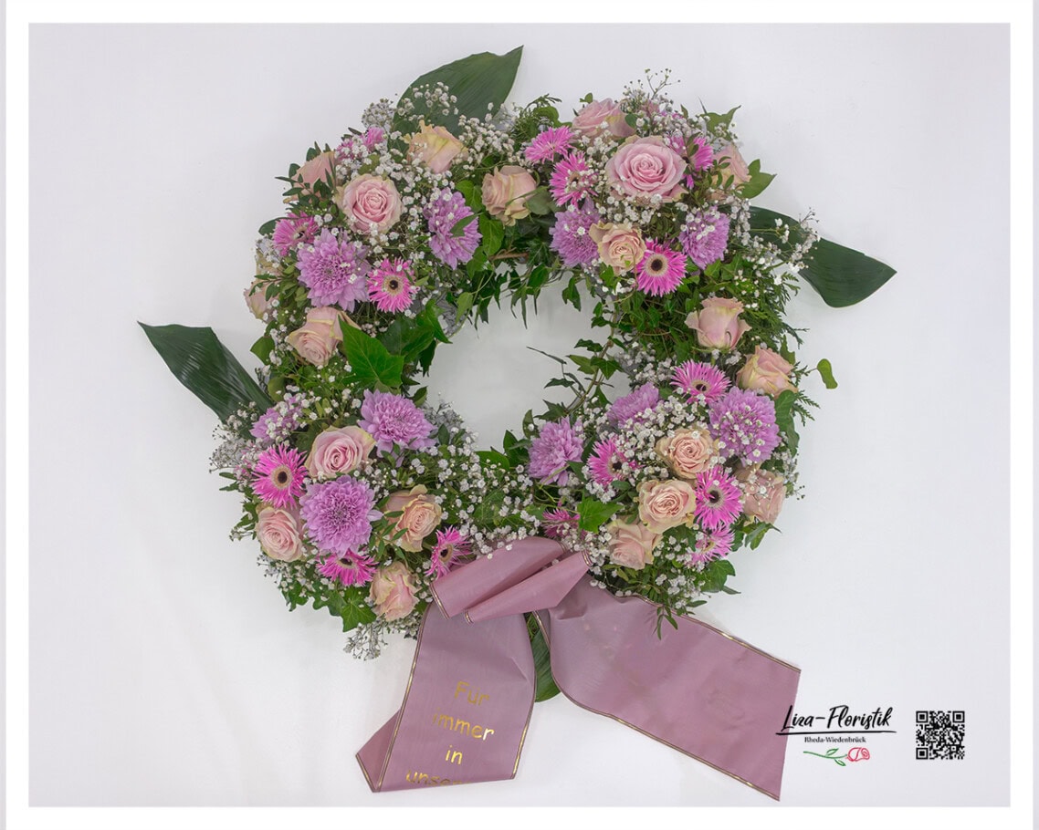 Trauerkranz mit zartrosa Rosen, pinken Gerbera, violetten Chrysanthemen und Schleierkraut, arrangiert auf einem grünen Blätterkranz mit Schleife.