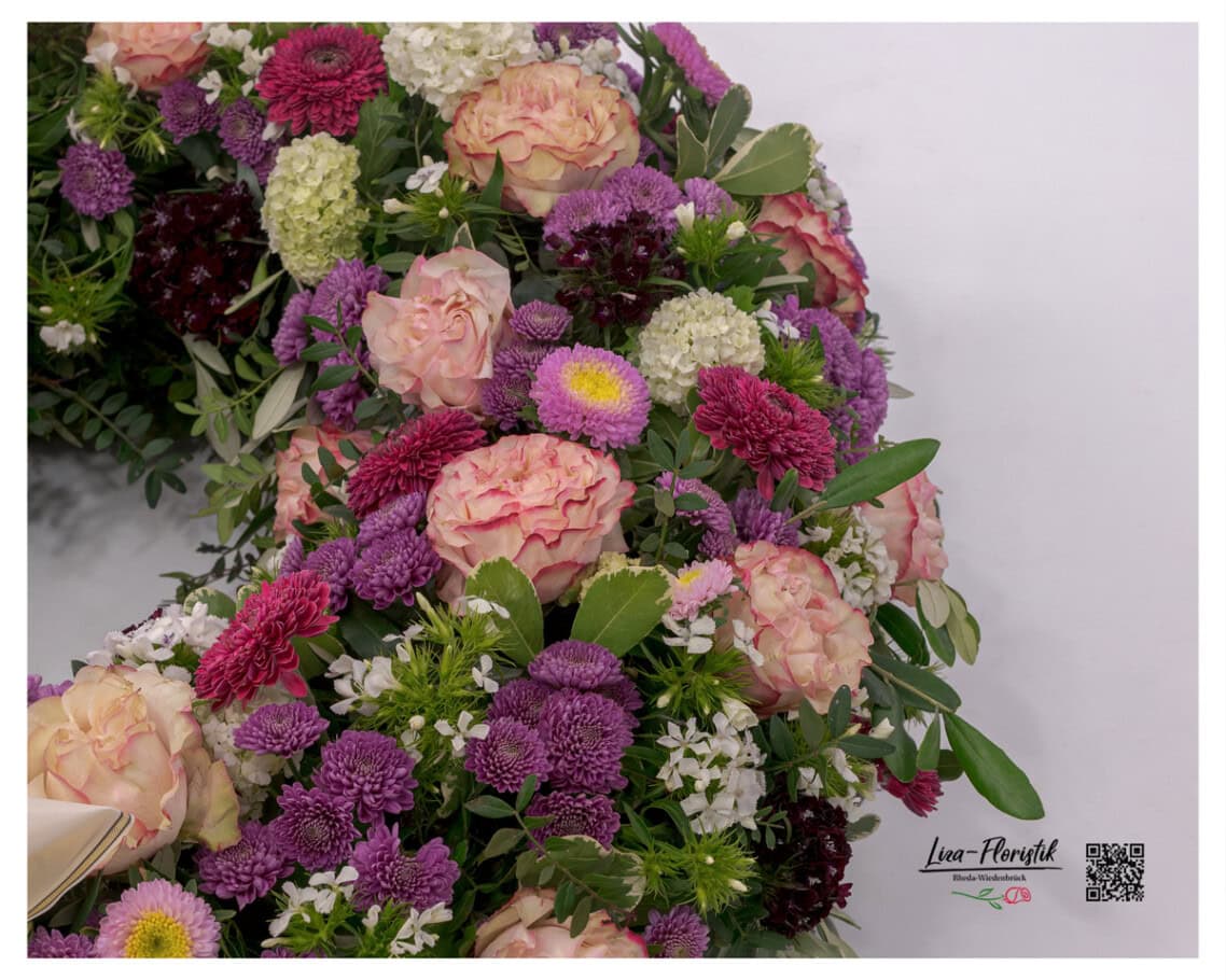 Draufsicht auf die dichte Blütenfülle des Kranzes – lachsfarbene Rosen, Bartnelken, lila Astern, Gerbera und Santini zeigen die handwerkliche Sorgfalt, mit der jede einzelne Blume gesetzt wurde.