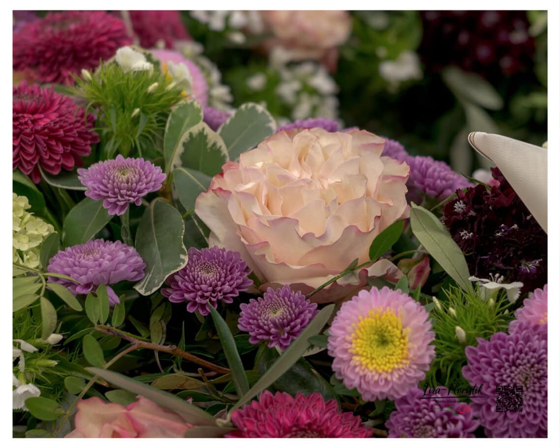 Cremeweiße Rose als Mittelpunkt, eingebettet in lila Astern, dunkles Santini und frisches Grün – die Farbkomposition des Kranzes aus nächster Nähe.