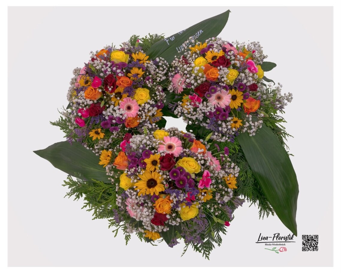 Trauerkranz in bunt mit drei Blütenarrangements – gelbe Sonnenblumen, orange und gelbe Premium Rosen, pinke Gerbera, dunkelrote Rosen, weißes Schleierkraut und violetter Statice, eingebettet in Thuja und Bananenblätter, handgefertigt von Liza-Floristik Rheda-Wiedenbrück